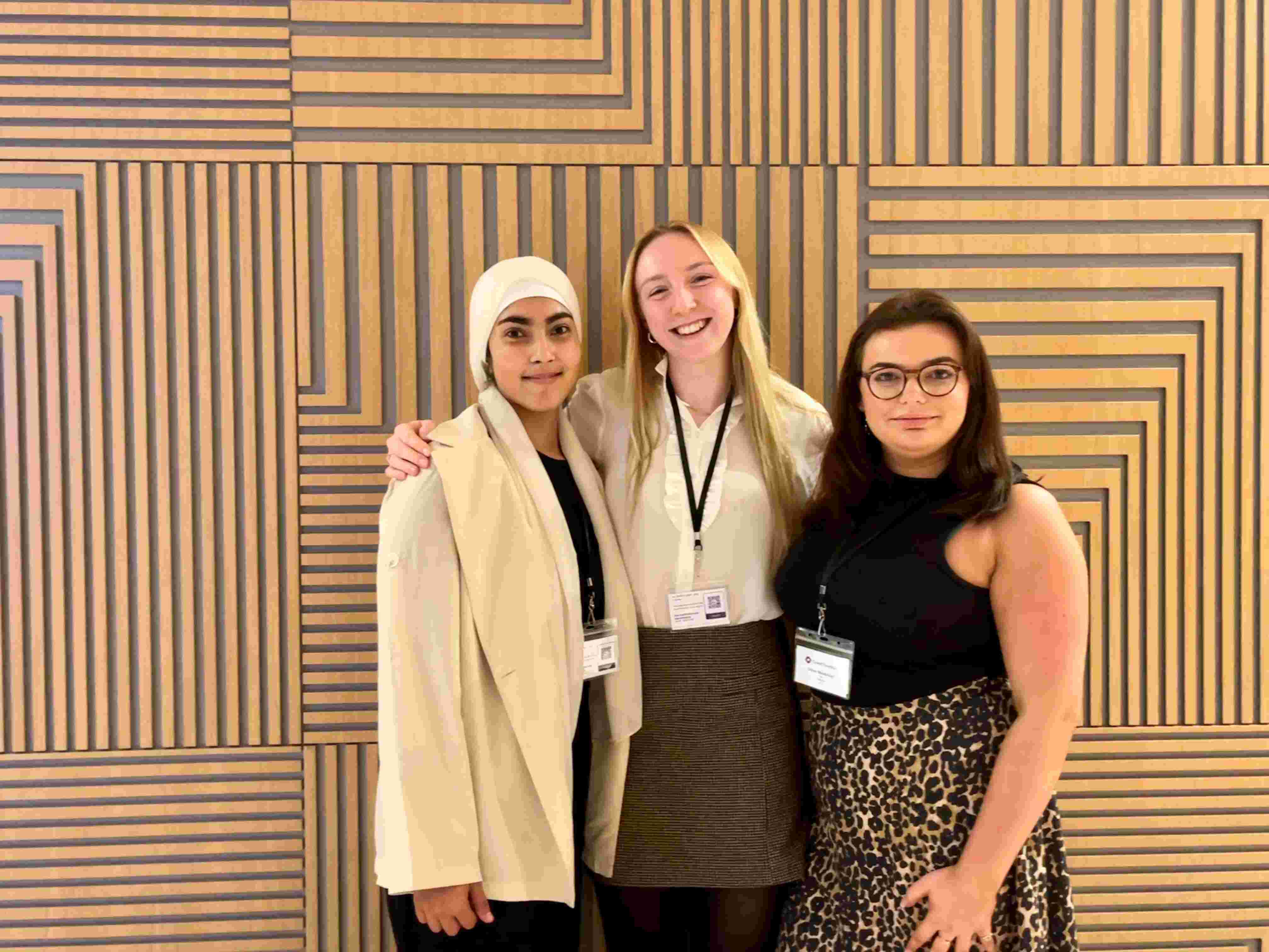 Three young women stand indoors against a beige geometric background, attending an event. They are all smiling at the camera and have their arms around each other.