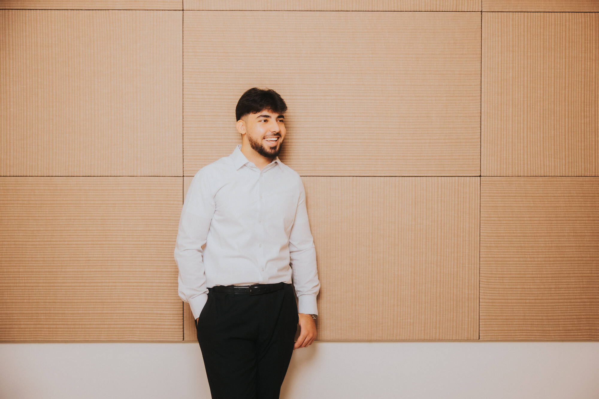 A young man with short dark brown hair is smiling to someone off camera. He is standing inside and is learning against a beige coloured wall. He is wearing a formal white shirt and black trousers.