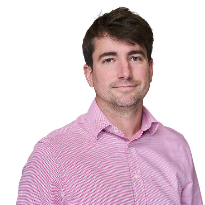 A man with short brown hair stands facing the camera with a smile, wearing a pink collared shirt. 