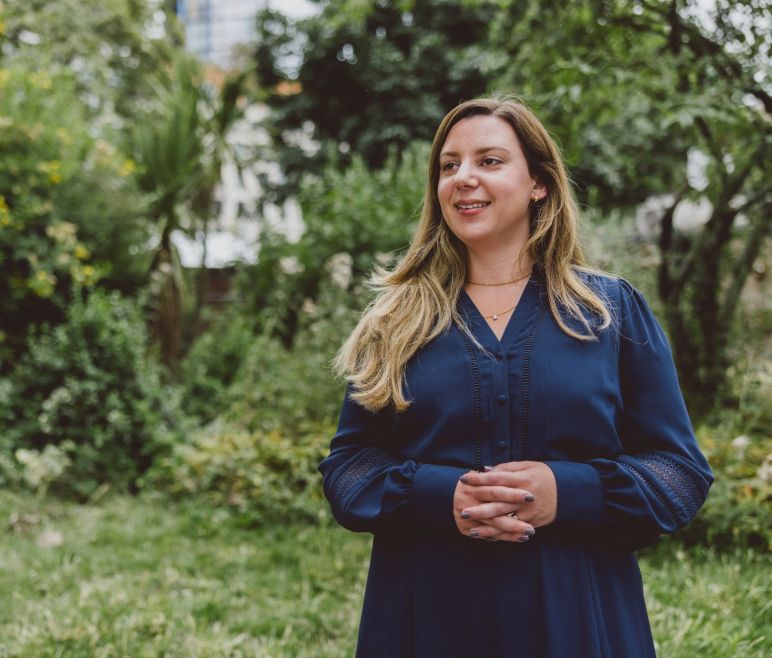 A woman stands in a garden wearing a blue dress, smiling while holding her hands together. The background is filled with green foliage, creating a serene atmosphere.