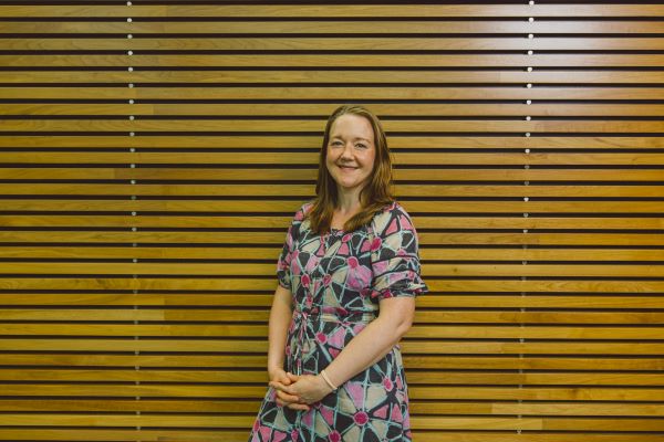 A lady is pictured standing in front of a wooden slat wall. She is smiling at the camera and wearing a grey dress with brightly coloured patterns.