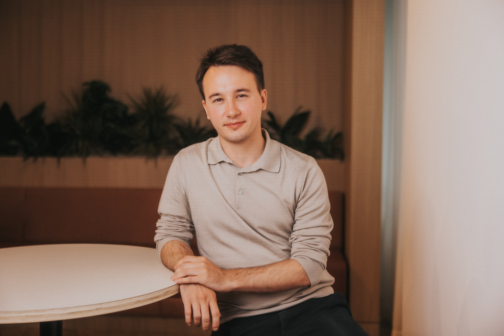 A young man with short blonde hair and a beige polo shit looks into the camera. He's sat in a chair with his arm resting on a desk, smiling.