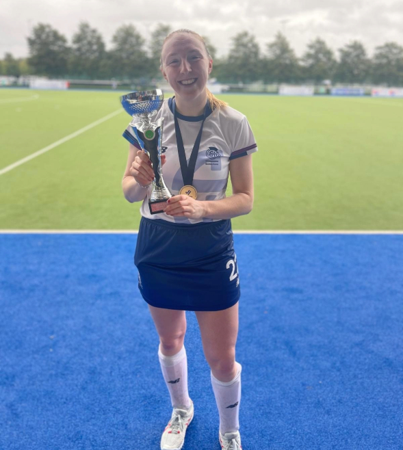 A young woman with blonde hair tied in a ponytail is smiling at the camera. She is standing on a hockey field outdoors and wearing a hockey uniform, holding a winners trophy. 
