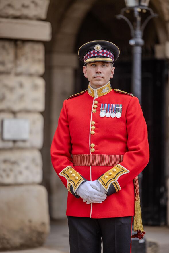 A man in a red ceremonial military uniform stands with hands clasped.