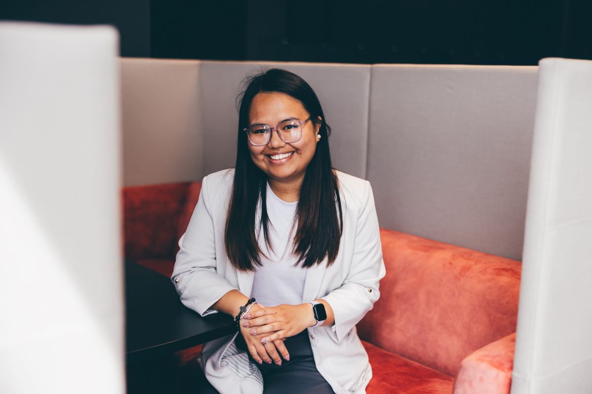 A Filipino woman is is pictured wearing a beige suit jacket and grey trousers. She is sitting in an office booth seat, with is coral. 