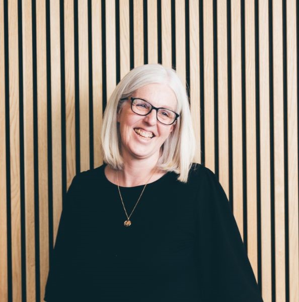 A woman with shoulder length grey hair is stood facing the camera, pictured waist upwards and is smiling broadly wearing a black top. 