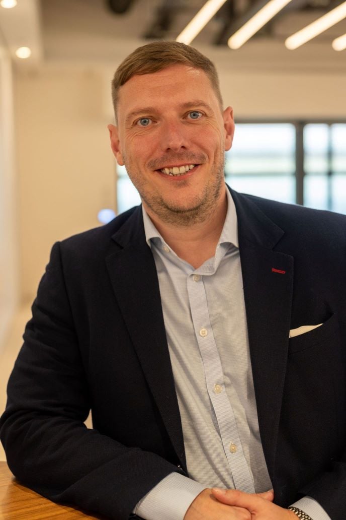 A man is pictured leaning against a table, from the waist up. He is wearing a light shirt and a dark jacket, with short brown hair and is looking directly at the camera. 