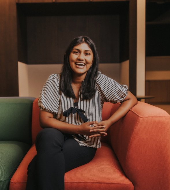 A young Asian woman sits inside in a comfortable looking orange and green chair. She is looking directly at the camera with a big smile. She has long black hair, a black and white striped blouse tied with bows at the front, and black trousers.