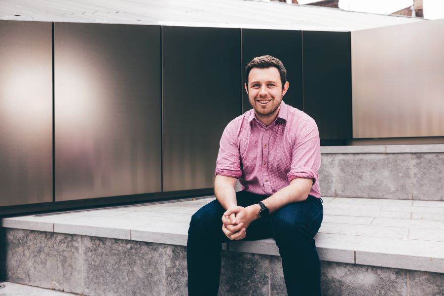A young man is pictured wearing a pink shirt. He is sat on steps in a courtyard area. 