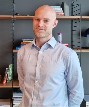 A man is smiling at the camera in an office, with a bookcase behind him. He is bald and wearing a light blue shirt.