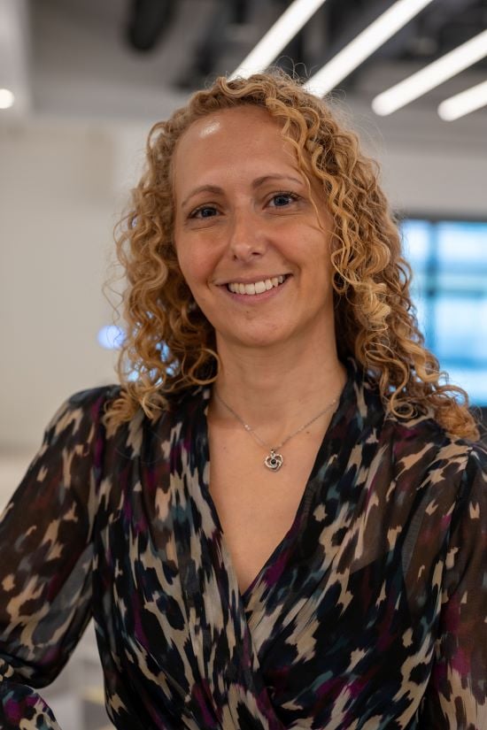 A woman with curly blonde hair is smiling at the camera in a close up shot of her head and shoulders. She's wearing a black, cream, red, and blue coloured blouse and silver necklace.