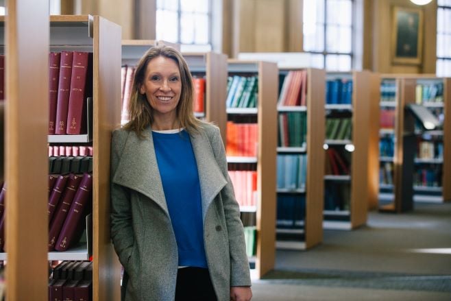 A woman with medium-length blonde hair, a grey coat and a blue top is standing in a library in a head and shoulders shot, smiling to the camera.