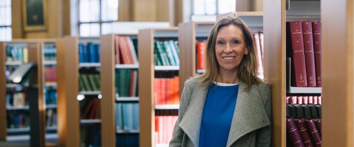 A woman is stood in a library, with rows of books behind her. She is looking straight at the camera, smiling, wearing a grey overcoat and a vibrant blue top. 