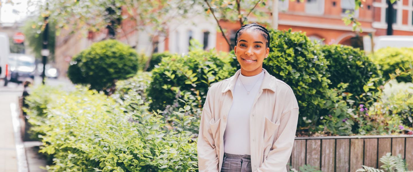 A young woman is pictured leaning against a raised flower bed in a city space with dark-brick buildings in the background. She is looking directly at the camera and smiling, wearing grey tailored trousers and a pale cream top and shirt.