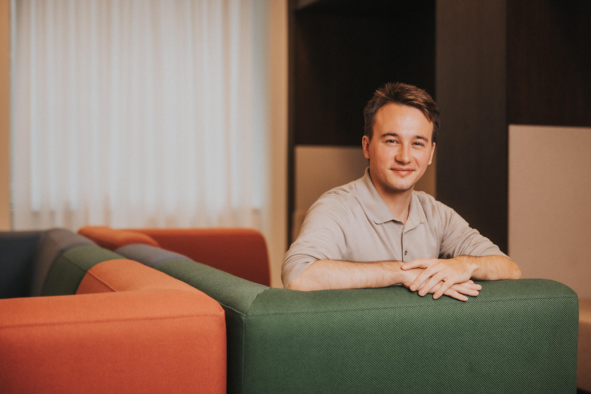 A young man with short blonde hair and a beige polo shirt looks into the camera. He's sat in a green chair next to an orange chair.