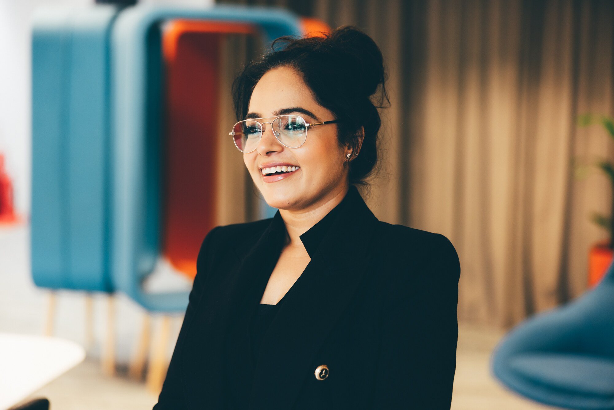 An Indian women is pictured in a head and shoulders shot, wearing a suit jacket. She has her tied back, and is wearing glasses looking off camera to the right. She is in a relaxed office environment.