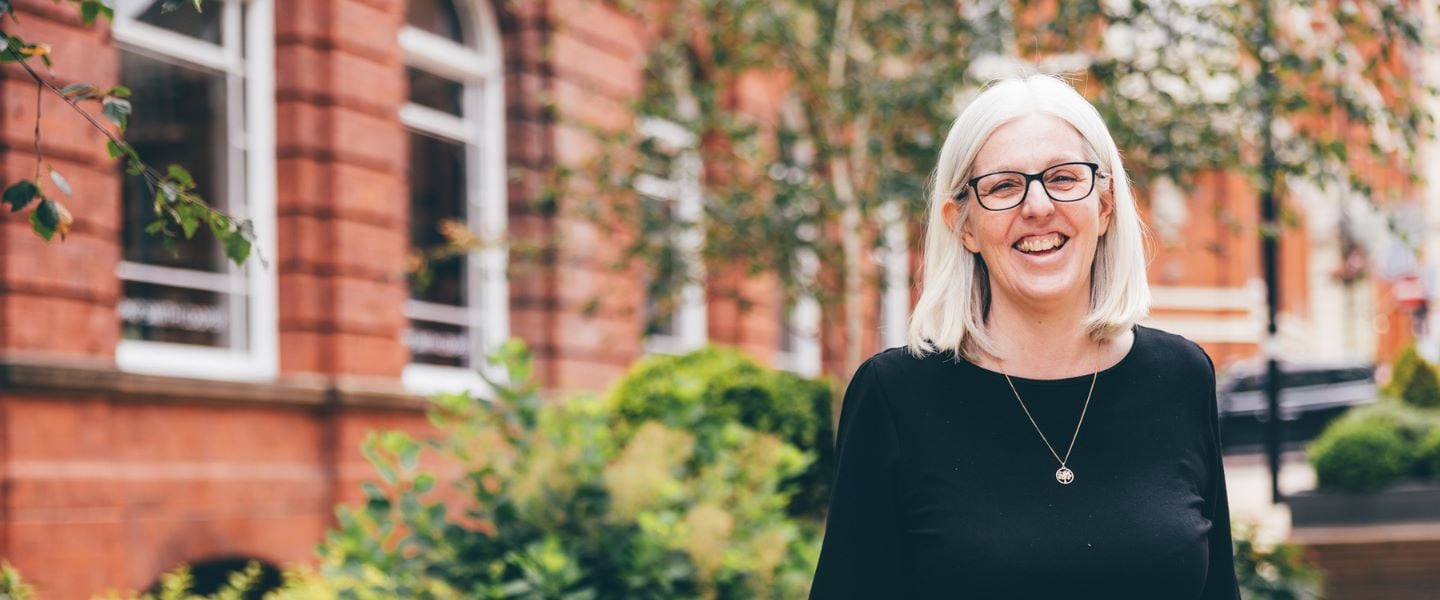 A woman with shoulder length grey hair is standing in a green city centre space, with buildings in the background. She is stood facing the camera, pictured waist upwards and is smiling broadly wearing a black top.