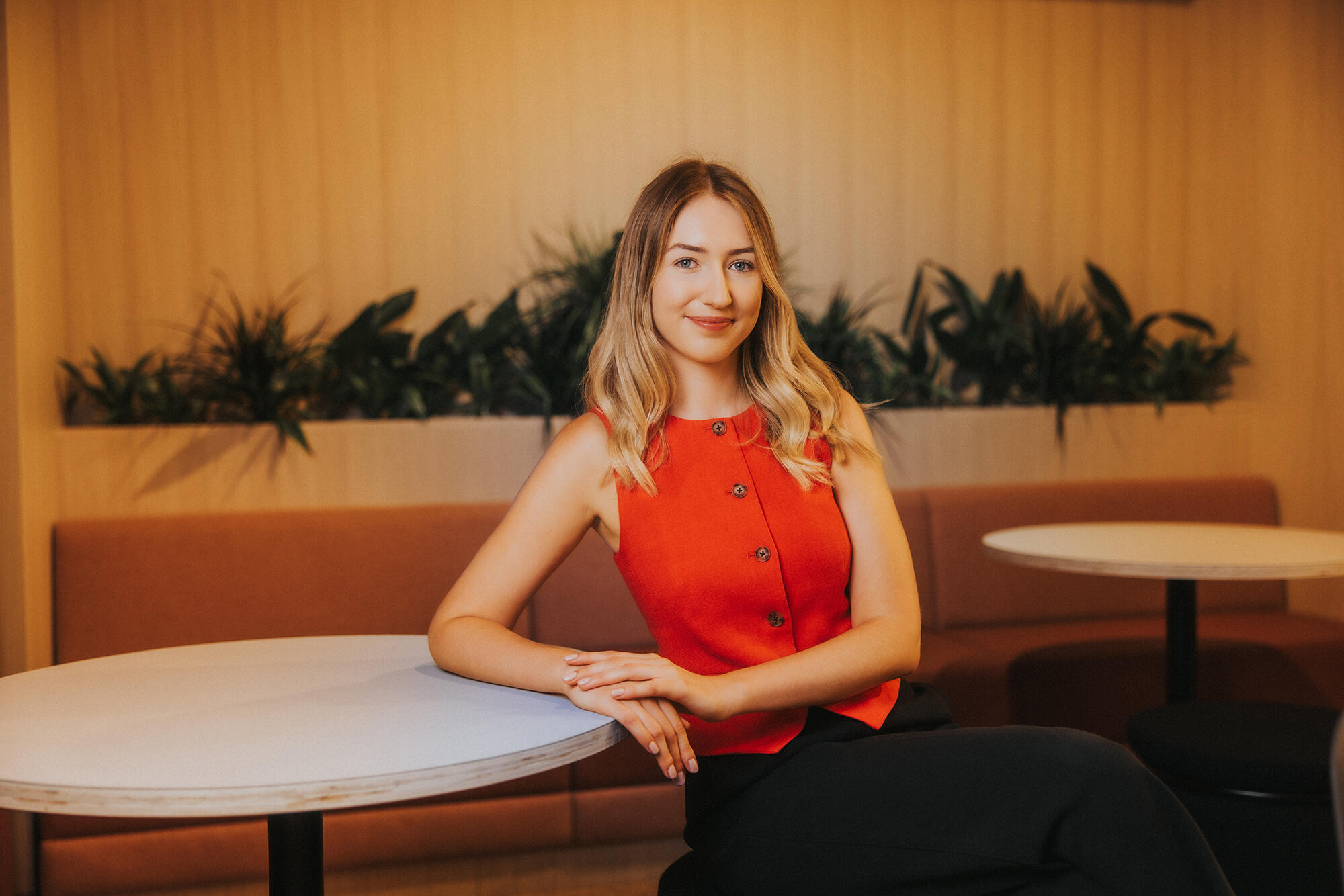 A young woman smiles and looks into the camera. She is sitting down with her elbow resting on a table beside her, with foliage in the background. She has long blonde hair and is wearing a red buttoned-up vest top.