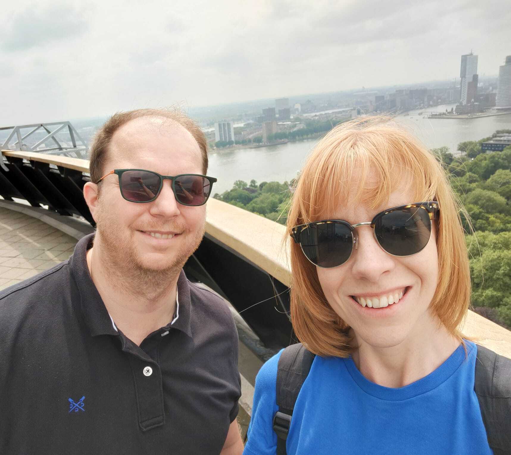A middle aged man and woman stand in front of a scenic backdrop, smiling at the camera. Both are wearing sunglasses. The man is wearing a grey polo shirt and the woman is wearing a blue t-shirt.
