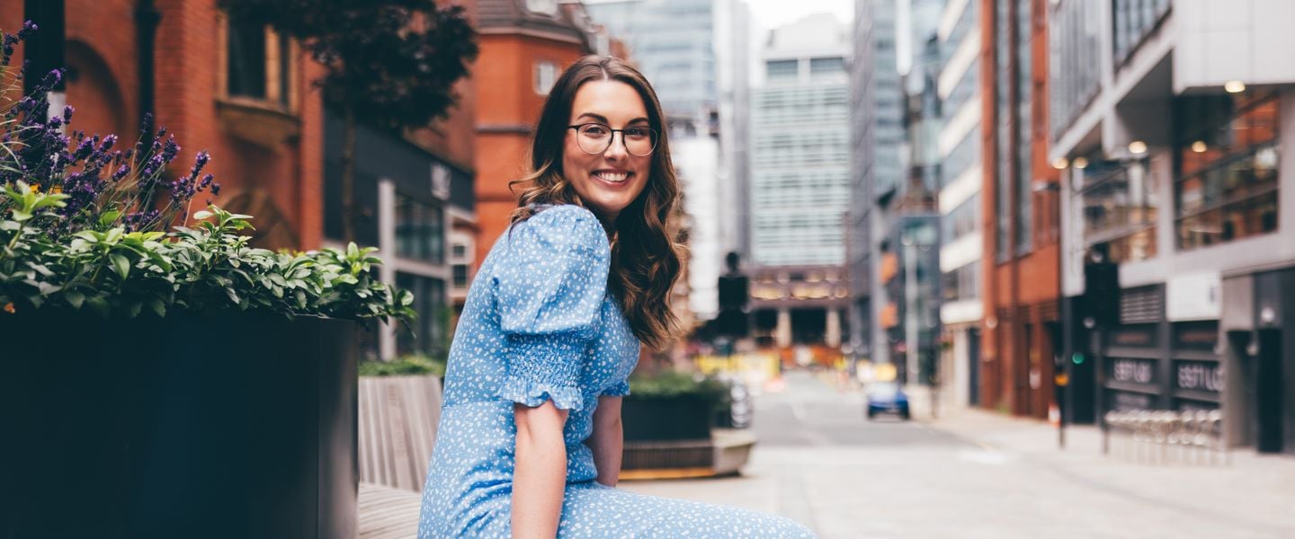 A young woman with long wavy brown hair is sat smiling at the camera, wearing a pale blue and white summer dress. She is wearing glasses and is pictured sitting outside on a bench in a city centre.