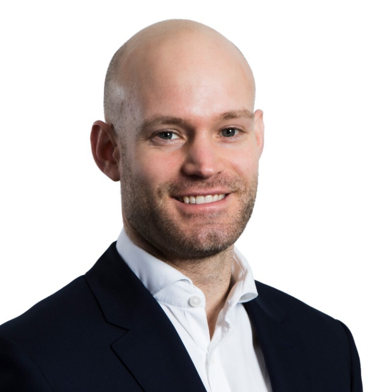 A man is smiling at the camera in a professional head and shoulders shot. He is bald and wearing a black suit blazer with a white shirt. 