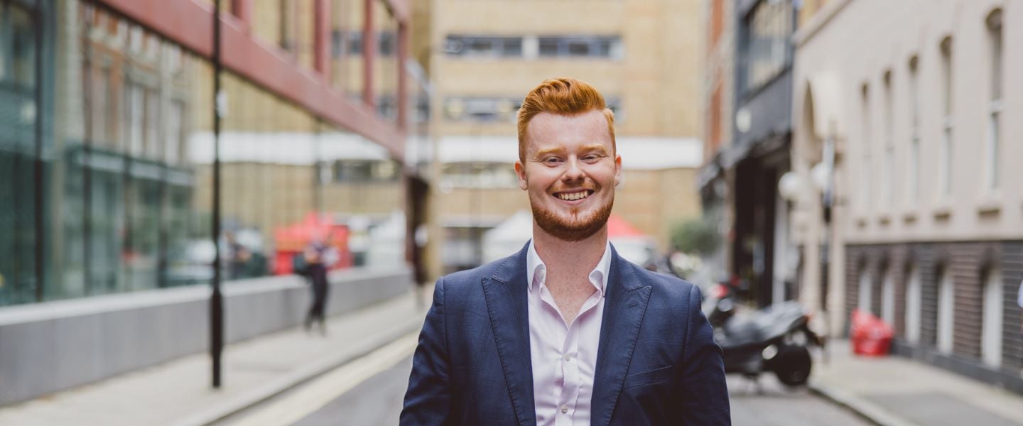 A young man with ginger hair is smiling whilst walking towards the camera. He is wearing a pale pink shirt and a grey suit jacket. 