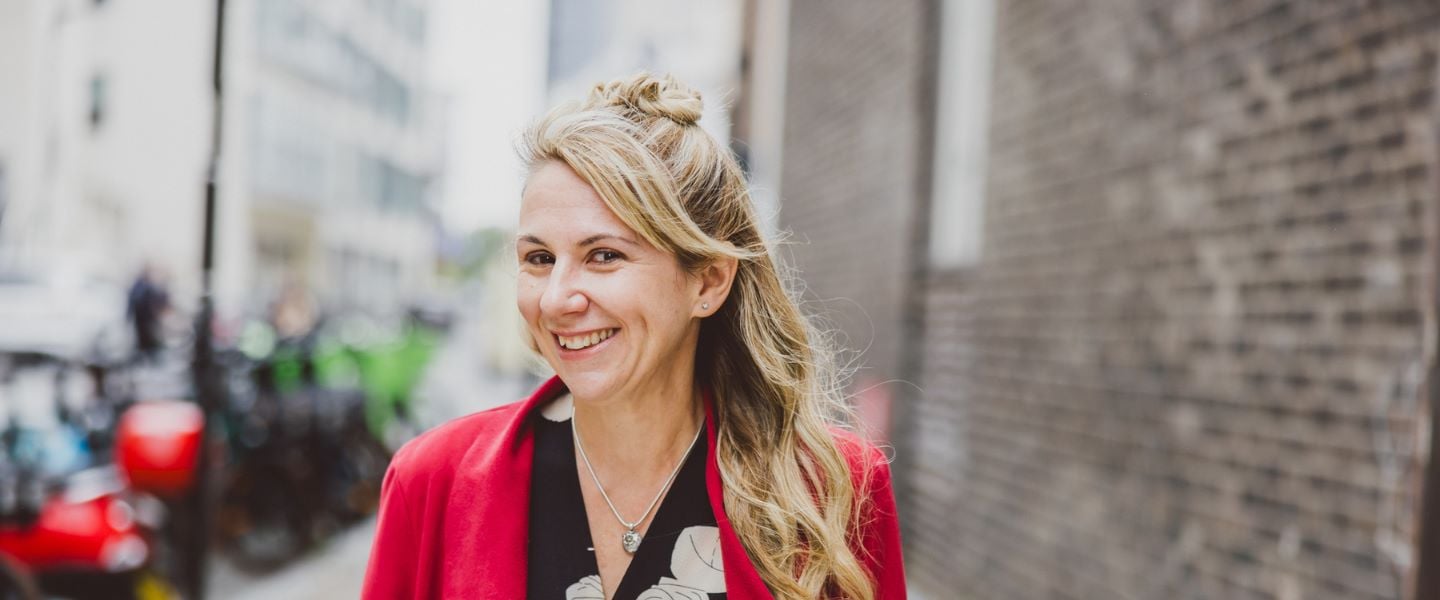 A woman with long blond hair is pictured walking towards the camera, smiling. She is wearing a black and white top with a bright red jacket.