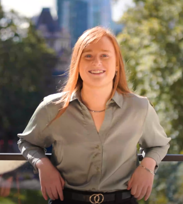 A young woman with long red hair is wearing a green silk shirt. She's standing outdoors on a sunny day, leaning against he balcony behind her, and smiling broadly at the camera.