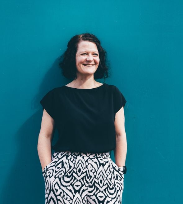 A woman with shoulder length brown hair pictured wearing a black top, in front of a bright teal wall. 