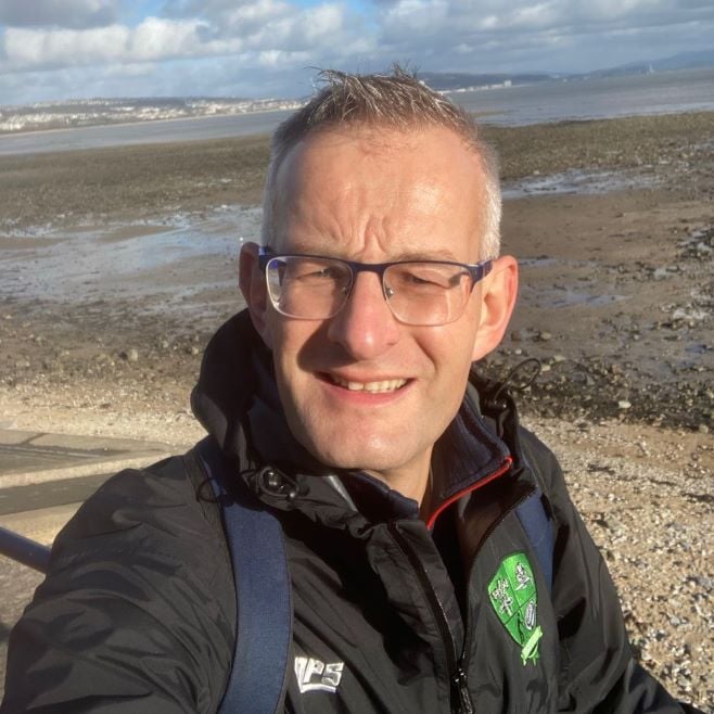 A man is photographed, with slightly grey hair, taking a selfie on a beach. 