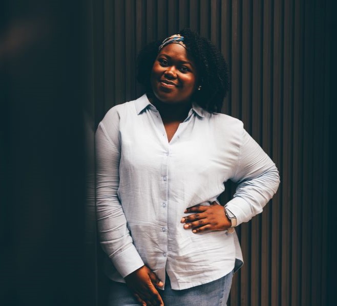 A woman is pictured in an standing in front of black glass wall, wearing a pale blue shirt and white trousers. She is smiling at the camera with a hand on her hip.