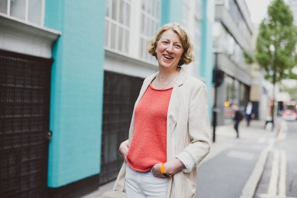 A woman is with short cropped hair, pictured smiling at the camera. She is wearing a coral top and beige jacket, standing in front of a building with a bright teal brick wall and grey shutters.