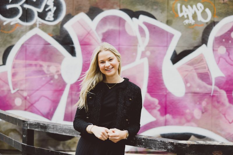 A young woman with long blond hair is photographed wearing a black top, stood against a fence in front of bright pink graffiti. 