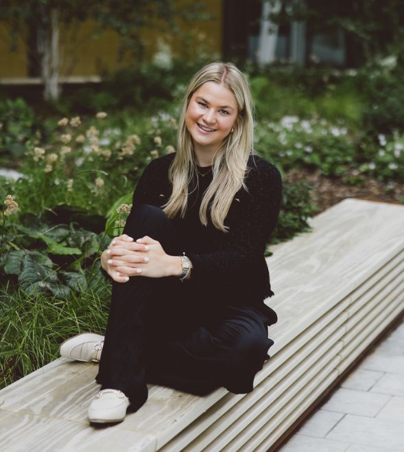 A young woman with long blond hair is photographed wearing a black top, sat on a bench outdoors and smiling at the camera.