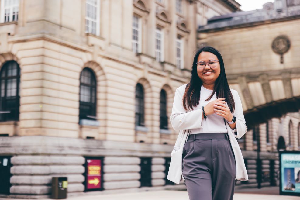 A Filipino woman is is pictured wearing a beige suit jacket and grey trousers. She is walking towards the camera smiling.