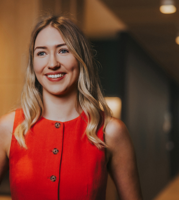 A young woman smiles and looks into the distance. She has long blonde hair and is wearing a red buttoned-up vest top.