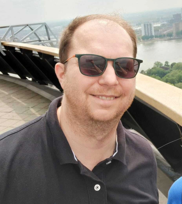 A middle aged man is stood in front of a scenic backdrop, smiling at the camera. He is wearing sunglasses and a grey polo shirt.