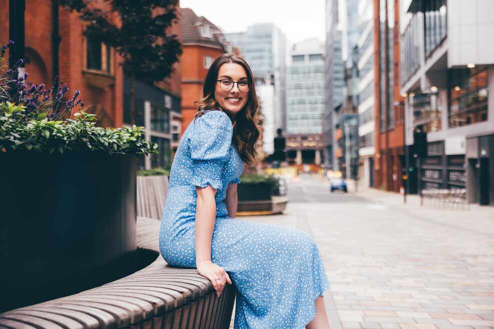 A woman with long brown hair and glasses is smiling to the camera, wearing a long blue dress with white flecks. She is sat on a bench outside on an empty street with flowers next to her.