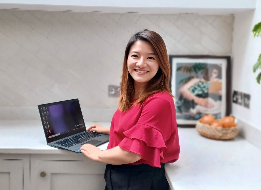 A woman with shoulder length brown hair is photographer in a kitchen setting, her laptop on the worktop. She is turned towards the camera, and wearing a bright crimson top. 