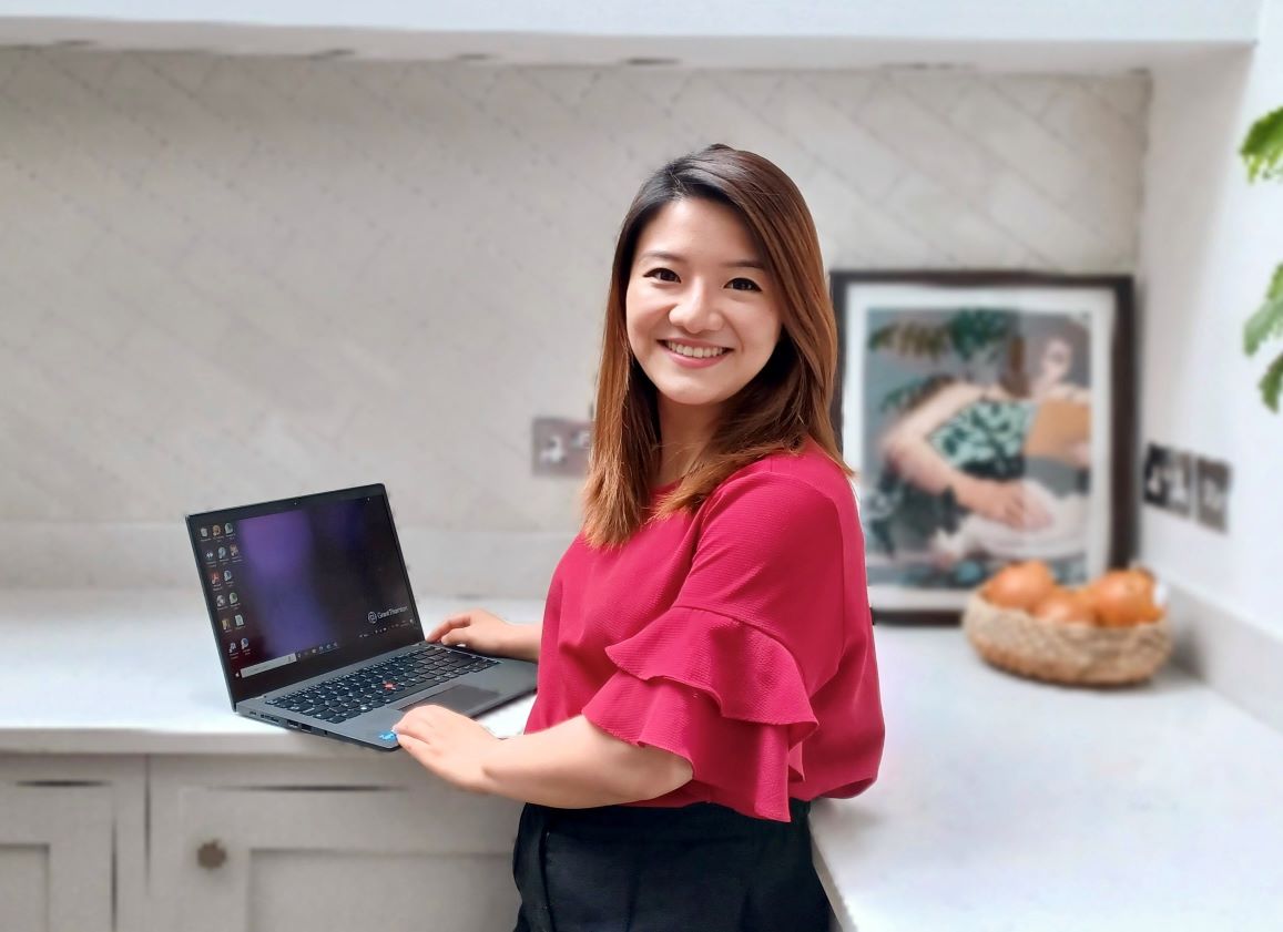 A woman with shoulder length brown hair is photographer in a kitchen setting, her laptop on the worktop. She is turned towards the camera, and wearing a bright crimson top. 