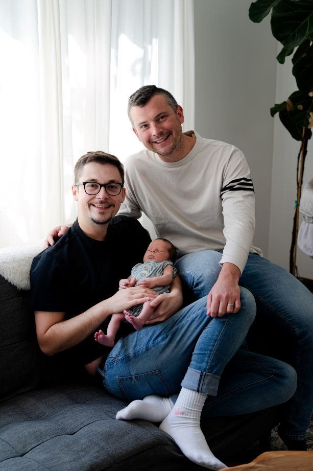Two men are photographed sitting on a sofa, with their newborn child.