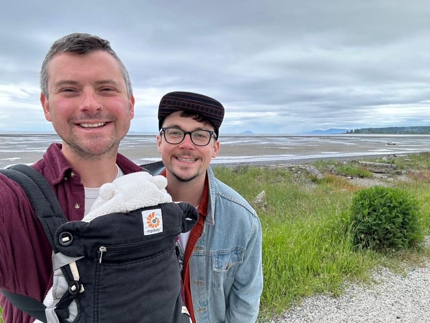 Two men are photographed on a coastal path, with their newborn child in a carrier.