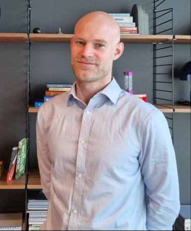 A man is smiling at the camera in an office, with a bookcase behind him. He is bald and wearing a light blue shirt.