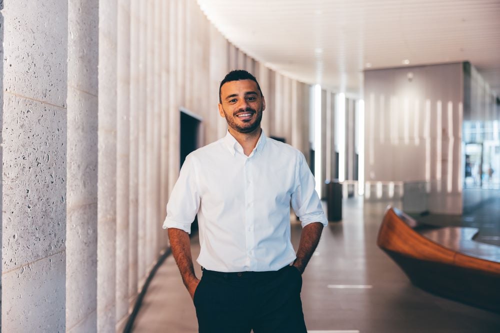 A man wearing a white shirt if pictured in a light and airy office reception area with concrete walls. 