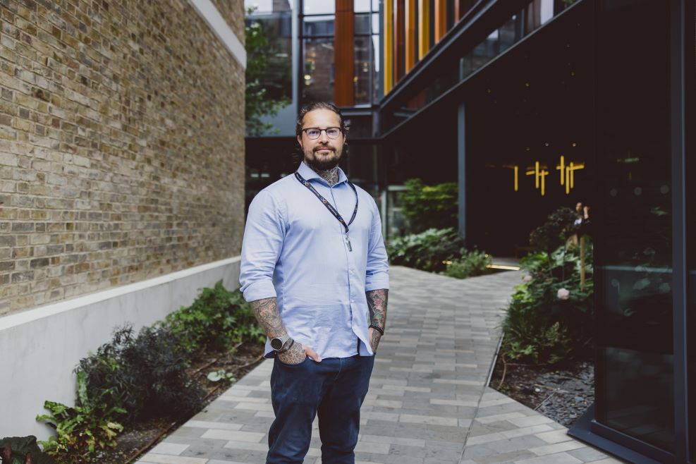 A man with a beard, glasses, and tattoos on his arms and neck is standing in front of a building. He is wearing a blue shirt.