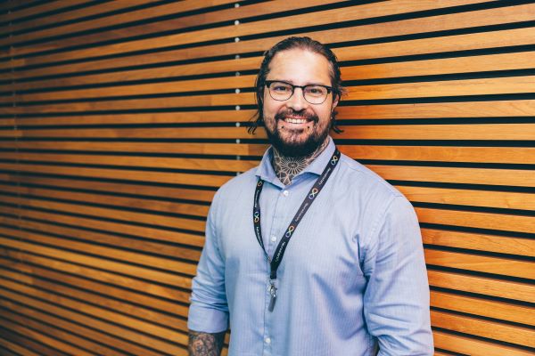 A man with a beard, glasses, and tattoos on his arms and neck is standing in front of a wooden backdrop, smiling. He is wearing a blue shirt and is photographed from the waist up.