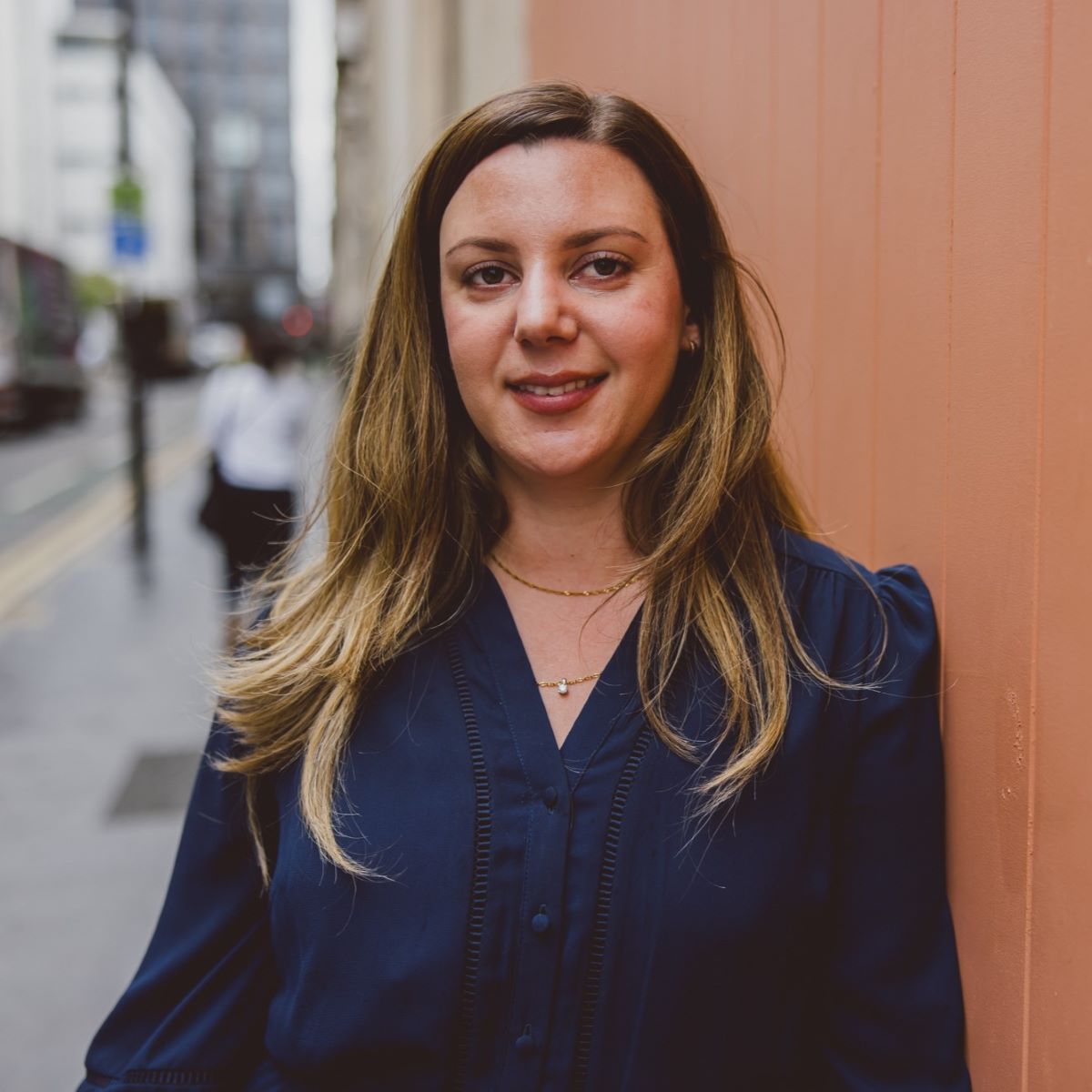 A woman with long brown hair is wearing a navy blue dress and pictured next to a peach door to her right, with a street behind her. 
