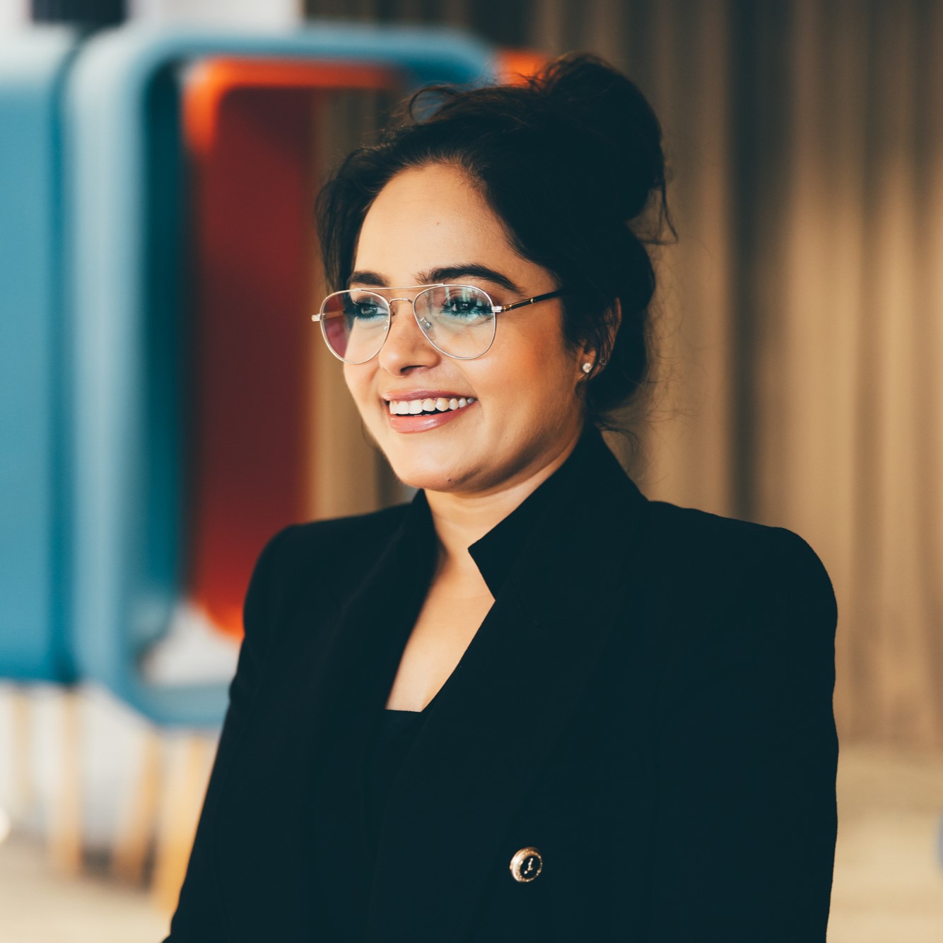A young woman is pictured wearing a black suit and glasses. She is looking slightly off camera, smiling. 