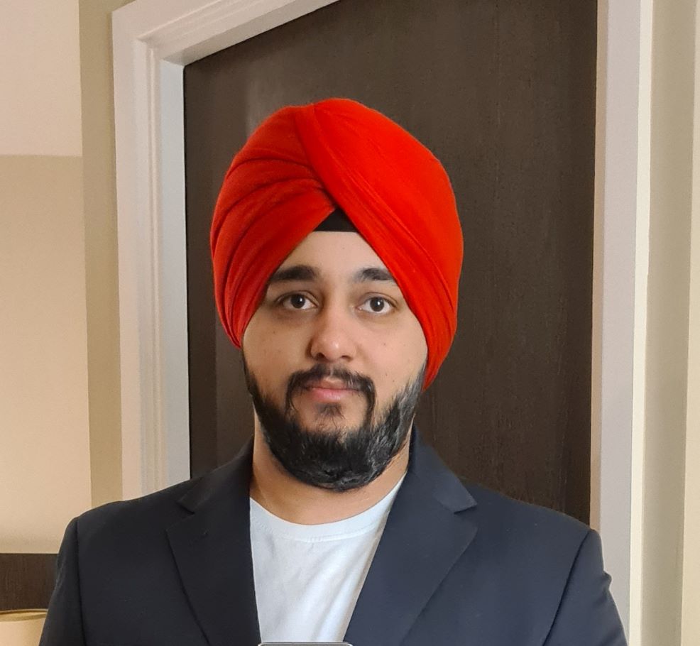 A man with a black beard wearing a bright red turban and a dark suit stands indoors. 